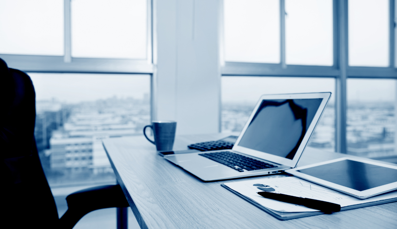 A blue washed out photograph of an vacated desk with open laptop papers and coffee mug next to a window with a view over city